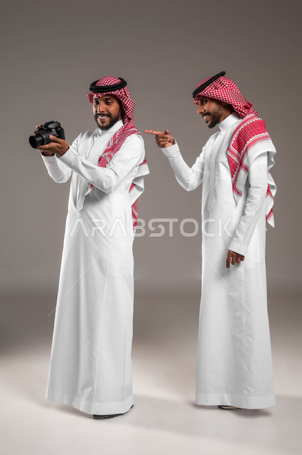 توثيق اللحظات السعيدة, استخدام تقنيات التصوير المبتكرة, الشغف بممارسة الهوايات مختلفة, بورتريه لتوءم شباب عربيين خليجين سعوديين يرتديان الثوب التقليدي والشماغ يستخدمان كاميرا فوتوغرافية عالية الدقة, صورة الجسم بالطول الكامل, خلفية رمادية