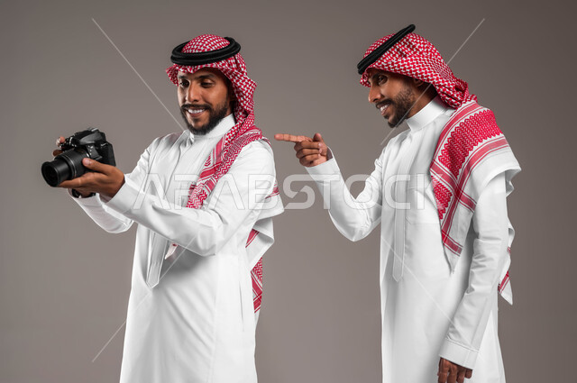اختيار الصور المناسبة وحفظها, الشغف بممارسة هوايات مختلفة, توثيق اللحظات السعيدة, بورتريه مقرب لتوءم شباب عربيين خليجين سعوديين يرتديان الثوب التقليدي والشماغ يستخدمان كاميرا فوتوغرافية عالية الدقة, استخدام تقنيات التصوير المبتكرة, خلفية رمادية
