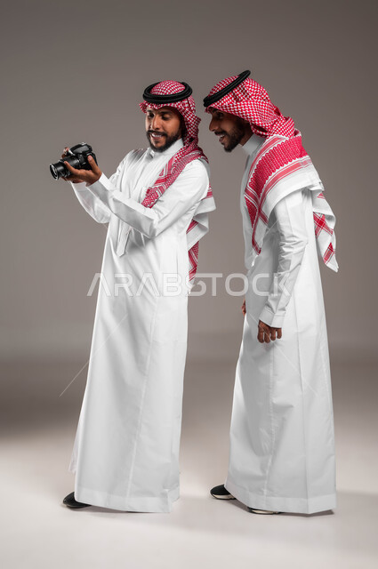 Using innovative photography techniques, passion for practicing different hobbies, documenting happy moments, portrait of twin Saudi Gulf Arab men wearing traditional thobes and shemaghs, using a high-resolution photographic camera, full-length body photo, gray background