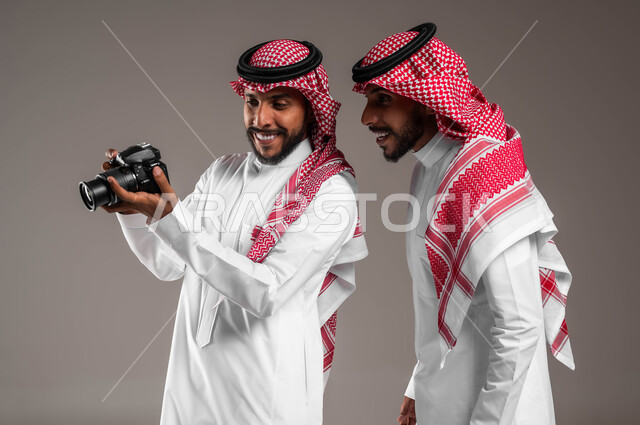 الشغف بممارسة هوايات مختلفة, توثيق اللحظات السعيدة, بورتريه مقرب لتوءم شباب عربيين خليجين سعوديين يرتديان الثوب التقليدي والشماغ يستخدمان كاميرا فوتوغرافية عالية الدقة, استخدام تقنيات التصوير المبتكرة, خلفية رمادية
