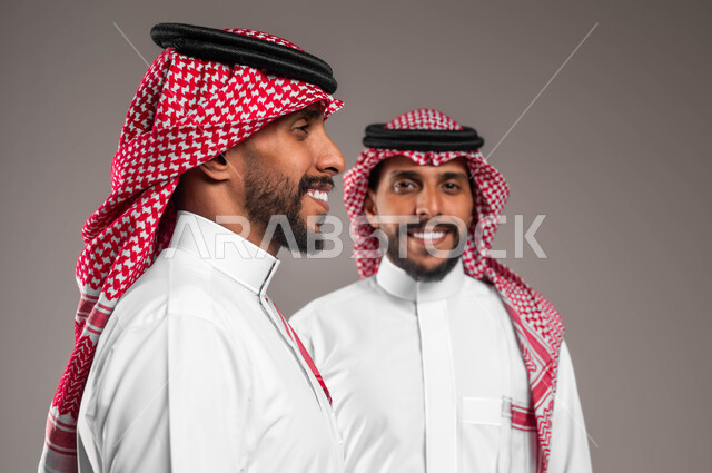 إيماءات وتعابير السرور والسعادة، التحديق إلى شيء ما بإيماءات التركيز، بورتريه مقرب لشابين توءم عربيين خليجيين سعوديين مبتسمين يرتديان الشماغ والثوب التقليدي أحدهما ينظر إلى الجانب  والآخر ينظر إلى الكاميرا، مفهوم التأمل والتخطيط، خلفية رمادية