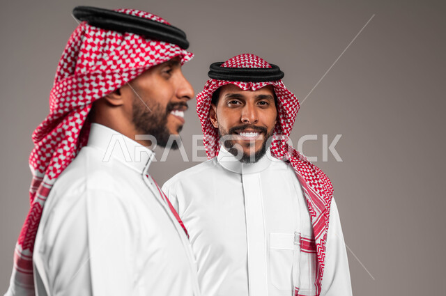 إيماءات وتعابير السرور والسعادة، التحديق إلى شيء ما بإيماءات التركيز، بورتريه مقرب لشابين توءم عربيين خليجيين سعوديين مبتسمين يرتديان الشماغ والثوب التقليدي أحدهما ينظر إلى الجانب  والآخر ينظر إلى الكاميرا، مفهوم التأمل والتخطيط، خلفية رمادية
