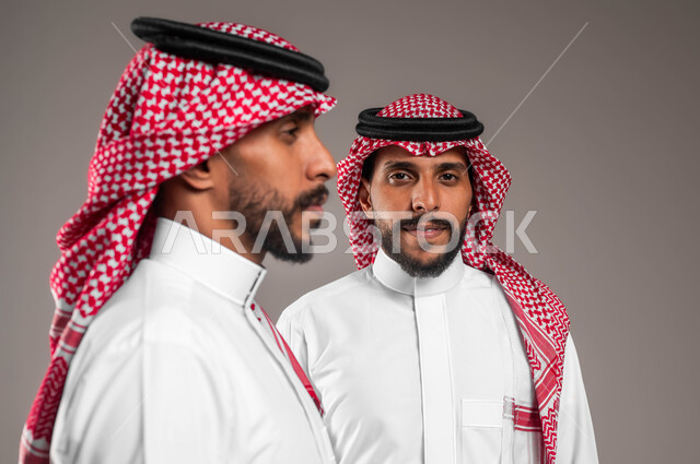 Staring at something with gestures of concentration, close-up portrait of two young Saudi Gulf Arab twins wearing a shemagh and traditional dress, one looking to the side and the other looking at the camera, concept of contemplation and planning, gray background