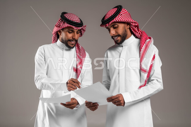 مفهوم العمل الجماعي، التعاون على تحقيق أهداف الشركة، حل مسائل الحسابات والأمور المالية، نقاشات وحوارات بين رجال الأعمال، بورتريه لشابين توءم عربيين خليجيين سعوديين يرتديان الثوب والشماغ التقليدي يحملان أوراق بيضاء، خلفية رمادية