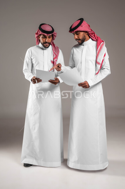 نقاشات وحوارات بين رجال الأعمال، مفهوم العمل الجماعي، التعاون على تحقيق أهداف الشركة، حل مسائل الحسابات والأمور المالية، بورتريه لشابين توءم عربيين خليجيين سعوديين يرتديان الثوب والشماغ التقليدي يحملان أوراق بيضاء بإيماءات التركيز، صورة لكامل الجسم، خلفية رمادية