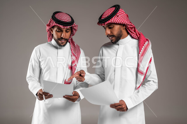 مفهوم العمل الجماعي، التعاون على تحقيق أهداف الشركة، حل مسائل الحسابات والأمور المالية، نقاشات وحوارات بين رجال الأعمال، بورتريه لشابين توءم عربيين خليجيين سعوديين يرتديان الثوب والشماغ التقليدي يحملان أوراق بيضاء، خلفية رمادية