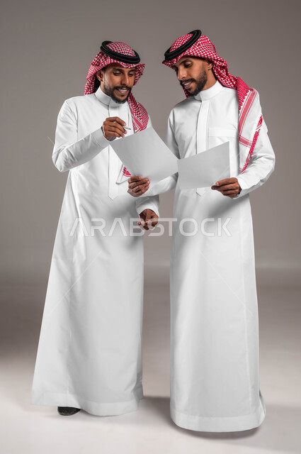 مفهوم العمل الجماعي، نقاشات وحوارات بين رجال الأعمال، التعاون على تحقيق أهداف الشركة، حل مسائل الحسابات والأمور المالية، بورتريه لشابين توءم عربيين خليجيين سعوديين يرتديان الثوب والشماغ التقليدي يحملان أوراق بيضاء بإيماءات التركيز، صورة لكامل الجسم، خلفية رمادية