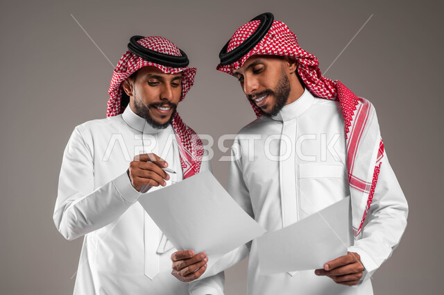 التعاون على تحقيق أهداف الشركة، حل مسائل الحسابات، نقاشات وحوارات بين زملاء العمل، بورتريه لشابين توءم عربيين خليجيين سعوديين يرتديان الثوب والشماغ التقليدي يحملان أوراق بيضاء، خلفية رمادية
