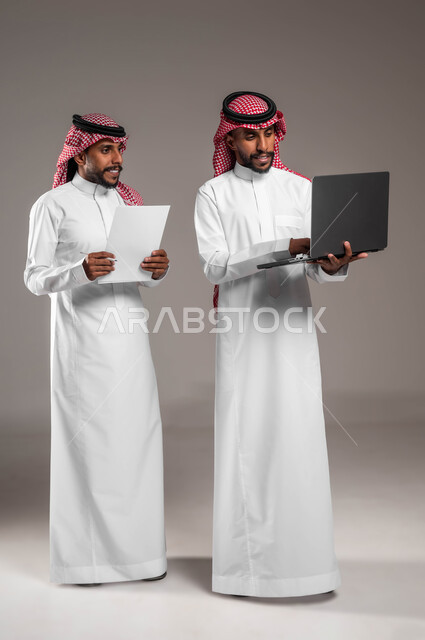 استخدام جهاز اللابتوب لإنجاز المهام، التعاون على تحقيق أهداف الشركة، الأجهزة والتقنيات الحديثة والمتطورة، بورتريه لشاب عربي خليجي سعودي يرتدي الثوب والشماغ التقليدي يقف بجانب أخيه التوءم، نقاشات وحوارات بين رجال الأعمال، صورة الجسم بالكامل، خلفية رمادية