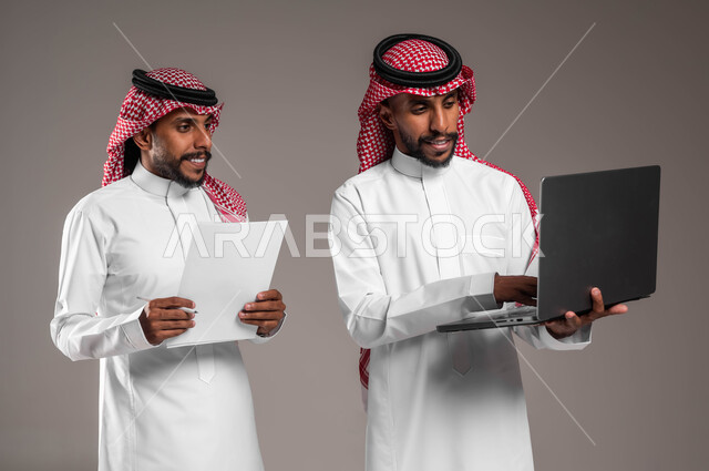 نقاشات وحوارات بين رجال الأعمال، استخدام جهاز اللابتوب لإنجاز المهام، التعاون على تحقيق أهداف الشركة، الأجهزة والتقنيات الحديثة والمتطورة، بورتريه لشاب عربي خليجي سعودي يرتدي الثوب والشماغ التقليدي يقف بجانب أخيه التوءم، خلفية رمادية