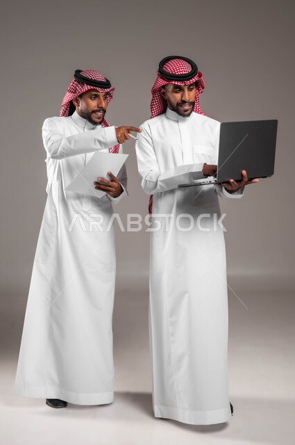 التعاون بين الزملاء في إنجاز المهام، استخدام الأجهزة والتقنيات الحديثة والمتطورة، بورتريه لشاب عربي خليجي سعودي يرتدي الثوب والشماغ التقليدي يقف بجانب أخيه التوءم ويشير إلى جهاز اللابتوب، نقاشات وحوارات بين رجال الأعمال، صورة الجسم بالكامل، خلفية رمادية