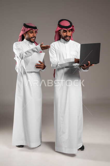 Cooperation between colleagues in completing tasks, using modern and advanced devices and technologies, a portrait of a Saudi Gulf Arab young man wearing the traditional thobe and shemagh standing next to his twin brother and pointing to the laptop, discussions and dialogues between businessmen, full body image, gray background