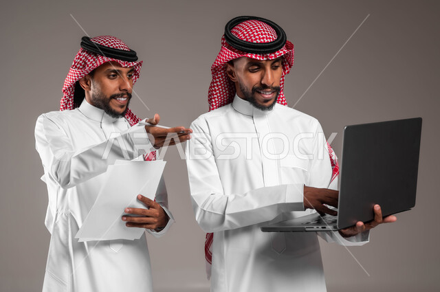 مفهوم التعاون والعمل الجماعي بين الزملاء في إنجاز المهام، استخدام الأجهزة والتقنيات الحديثة والمتطورة، بورتريه لشاب عربي خليجي سعودي يرتدي الثوب والشماغ التقليدييقف بجانب أخيه التوءم ويشير إلى جهاز اللابتوب، نقاشات وحوارات بين رجال الأعمال، خلفية رمادية