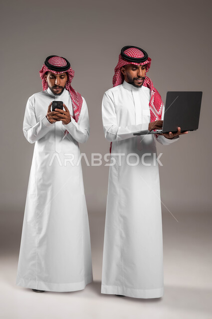 إيماءات الاندماج والتركيز أثناء استخدام الأجهزة والتقنيات الحديثة والمتطورة، بورتريه لشابين توءم عربيين خليجيين سعوديين يرتديان الثوب والشماغ التقليدي يحملان جهاز اللابتوب والهاتف المحمول، صورة لكامل الجسم، خلفية رمادية
