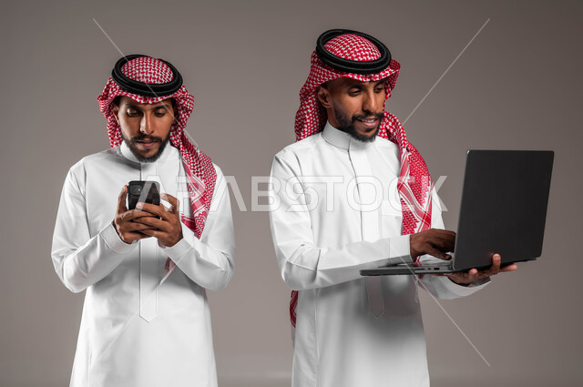 Gestures of integration and focus, using modern and advanced devices and technologies, seeing something through the computer, a portrait of two young Saudi Gulf Arab twins wearing the traditional thobe and shemagh, holding a laptop and mobile phone, gray background.