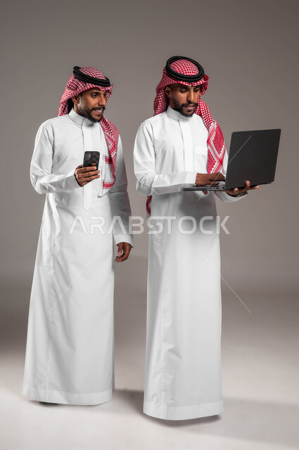 إيماءات الاندماج والتركيز أثناء استخدام الأجهزة والتقنيات الحديثة والمتطورة، بورتريه لشابين توءم عربيين خليجيين سعوديين يرتديان الثوب والشماغ التقليدي يحملان جهاز اللابتوب والهاتف المحمول، صورة لكامل الجسم، خلفية رمادية