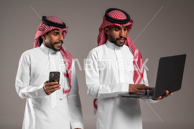 Gestures of integration and focus, using modern and advanced devices and technologies, seeing something through the computer, a portrait of two young Saudi Gulf Arab twins wearing the traditional thobe and shemagh, holding a laptop and mobile phone, gray background.