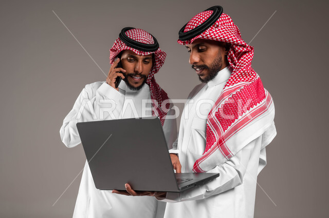 الأجهزة والتقنيات الحديثة والمتطورة، إجراء مكالمة هاتفية باستخدام الجوال، رؤية شيء ما عبر الحاسوب، بورتريه مقرب لشابين توءم عربيين خليجيين سعوديين يحملان جهاز اللابتوب والهاتف المحمول، خلفية رمادية