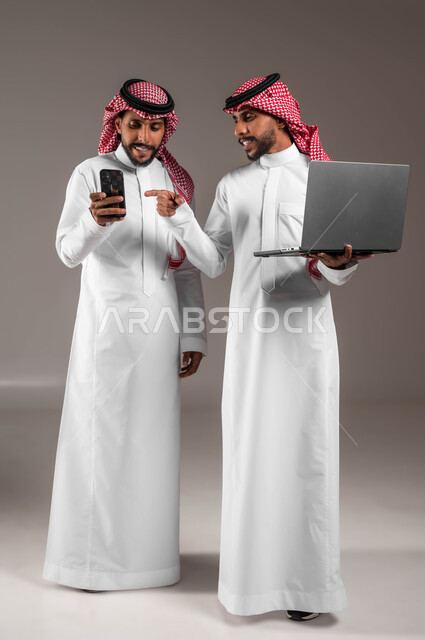 الإشارة بإصبع السبابة إلى الجوال، التعاون والمشاركة بين الأخوة لإنجاز المهام، بورتريه لشابين توءم عربيين خليجيين سعوديين مبتسميين يحملان جهاز اللابتوب والهاتف المحمول بإيماءات السرور والاعجاب بالتقنيات الحديثة، صورة الجسم بالكامل، خلفية رمادية