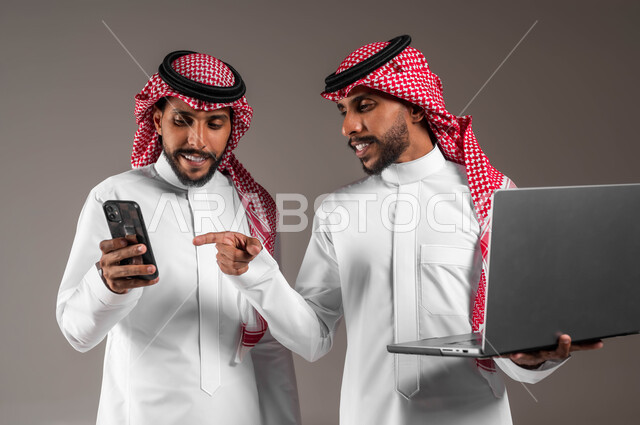 الإشارة بإصبع السبابة إلى الجوال، تعاون الأخوة لإنجاز المهام والأعمال، استخدام الأجهزة التقنية الحديثة والمتطورة, بورتريه مقرب لشابين توءم عربيين خليجيين سعوديين مبتسميين يحملان جهاز اللابتوب والهاتف المحمول بتعابير وايماءات السرور والاعجاب، خلفية رمادية
