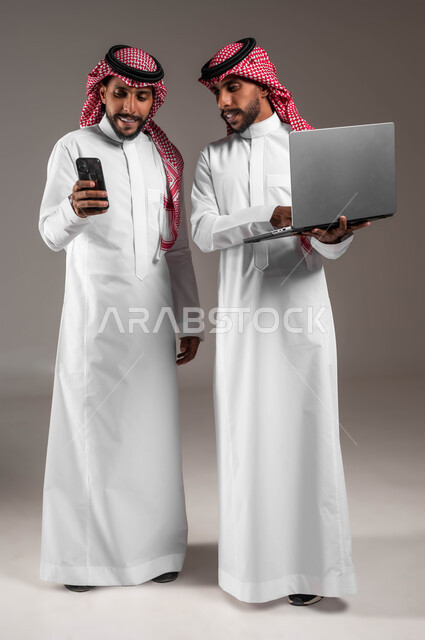 المشاركة والتعاون بين الأخوة لإنجاز المهام، بورتريه لشابين توءم عربيين خليجيين سعوديين مبتسميين يحملان جهاز اللابتوب والهاتف المحمول بإيماءات السرور والاعجاب بالتقنيات الحديثة، صورة الجسم بالكامل، خلفية رمادية