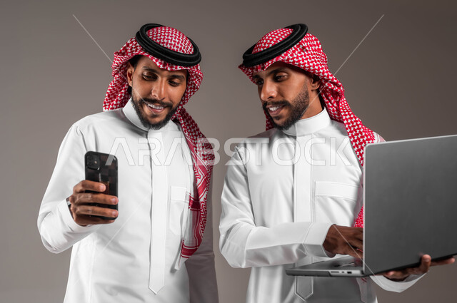 تعاون الأخوة لإنجاز المهام والأعمال، استخدام الأجهزة التقنية الحديثة والمتطورة, بورتريه مقرب لشابين توءم عربيين خليجيين سعوديين مبتسميين يحملان جهاز اللابتوب والهاتف المحمول، تعابير وايماءات السرور والاعجاب، خلفية رمادية