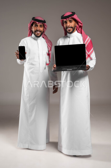 Expressions and gestures of pleasure and admiration, use of advanced modern technical devices, portrait of two smiling young Saudi Gulf Arab twins holding a laptop and a mobile phone, displaying a blank black screen, full-length body image, gray background