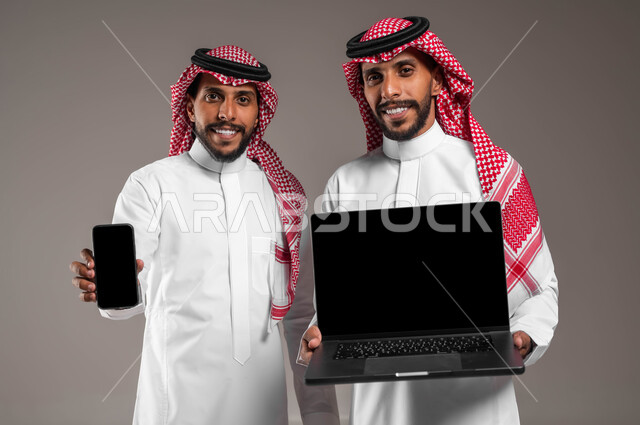 Using advanced modern technical devices, a close-up portrait of two smiling young Saudi Gulf Arab twins holding a laptop and a mobile phone, displaying a blank black screen, expressions and gestures of pleasure and admiration, a gray background.