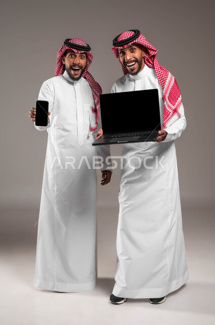 Expressions and gestures of shock and astonishment, use of advanced modern technical devices, a portrait of two young Saudi Gulf Arab twins holding a laptop and a mobile phone with a blank black screen, admiration and astonishment at something, full-length body image, gray background.