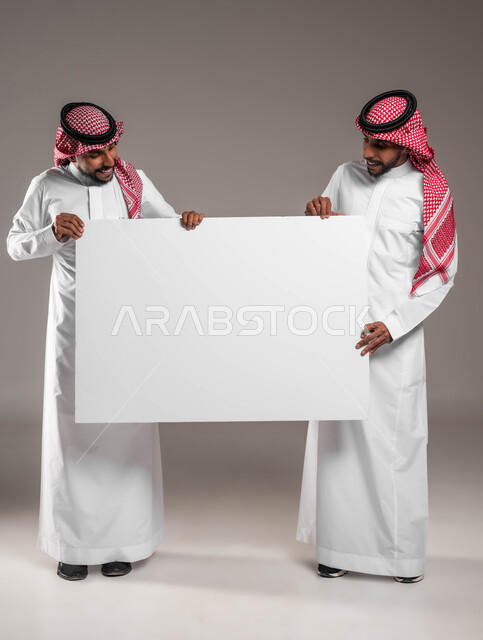 النظر الى موك اب مستطيل كبير بايماءات السرور والاعجاب، بورتريه لشابين توءم عربيين خليجيين سعوديين يرتديان الشماغ والثوب التقليدي يحملان  لوح اعلاني فارغ، إعلانات وعروض تسويقية، استخدام لوحات توضيحية ووسائل تعليمية، صورة الجسم بالكامل، خلفية رمادية