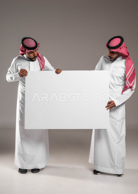 Looking at a large rectangular mock-up with gestures of astonishment and shock, a portrait of two young Saudi Gulf Arab twins wearing the shemagh and the traditional thobe, holding an empty billboard, advertisements and marketing offers, using explanatory panels and educational aids, full body image, gray background.