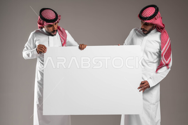 النظر الى موك اب مستطيل كبير بايماءات الذهول والصدمة، بورتريه مقرب لشابين توءم عربيين خليجيين سعوديين يرتديان الشماغ والثوب التقليدي يحملان لوح اعلاني فارغ، إعلانات وعروض تسويقية، استخدام لوحات توضيحية ووسائل تعليمية، خلفية رمادية