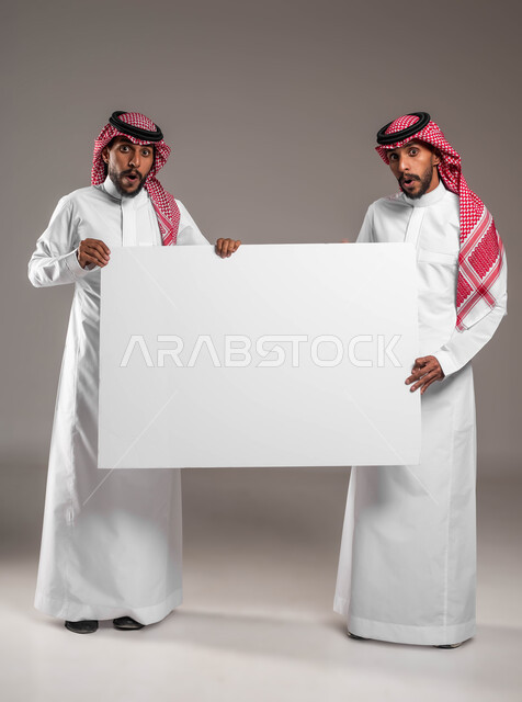 A large rectangular mockup, looking at the camera with expressions of astonishment and shock, a portrait of two young Saudi Gulf Arab twins wearing the shemagh and the traditional thobe, holding an empty billboard, advertisements and marketing offers, using explanatory panels and educational aids, full body image, gray background.