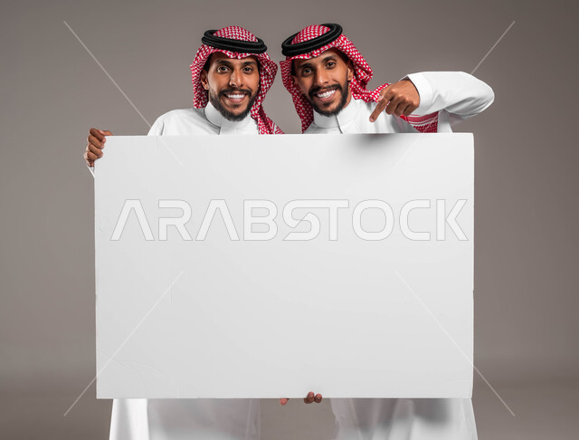 عروض واعلانات تسويقية، عرض واستخدام لوحات توضيحية ووسائل تعليمية، النظر الى الكاميرا والاشارة الى لوح اعلاني فارغ بتعابير السرور والاعجاب، بورتريه مقرب لشابين توءم عربيين خليجيين سعوديين يرتديان الشماغ والثوب التقليدي يحملان موك اب مستطيل كبير، خلفية رمادية