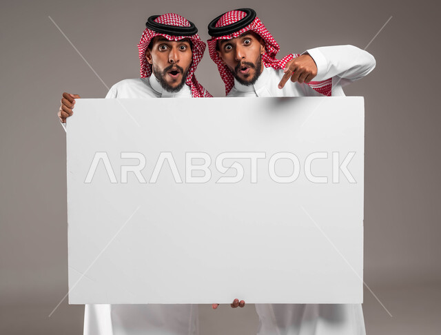 عرض واستخدام لوحات توضيحية ووسائل تعليمية، النظر الى الكاميرا والاشارة الى لوح اعلاني فارغ بتعابير الدهشة، بورتريه مقرب لشابين توءم عربيين خليجيين سعوديين يرتديان الشماغ والثوب التقليدي يحملان موك اب مستطيل كبير، إعلانات وعروض تسويقية،  خلفية رمادية