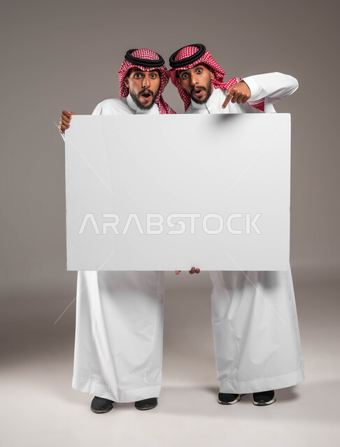 Looking at the camera and pointing to an empty billboard with expressions of amazement, a portrait of two young Saudi Gulf Arab twins wearing the shemagh and traditional thobe, holding a large rectangular mockup, advertisements and marketing presentations, use of explanatory panels and educational aids, full body portrait, gray background