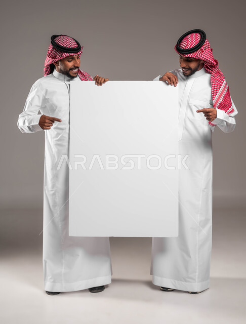 استخدام لوحات توضيحية ووسائل تعليمية، الاشارة الى لوح اعلاني فارغ بتعابير السرور والسعادة، إعلانات وعروض تسويقية، بورتريه لشابين توءم عربيين خليجيين سعوديين يرتديان الشماغ والثوب التقليدي يحملان موك اب مستطيل كبير ينظران اليه، صورة الجسم بالكامل، خلفية رمادية
