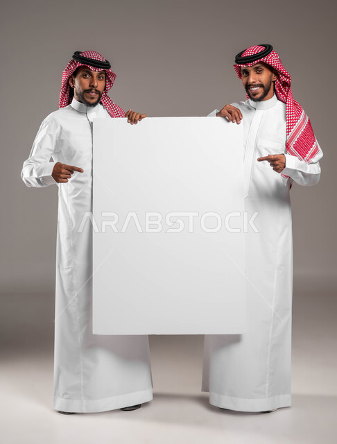 الاشارة الى لوح اعلاني فارغ بتعابير السرور والسعادة، استخدام لوحات توضيحية ووسائل تعليمية، إعلانات وعروض تسويقية، بورتريه لشابين توءم عربيين خليجيين سعوديين يرتديان الشماغ والثوب التقليدي يحملان موك اب مستطيل كبير،  صورة الجسم بالكامل، خلفية رمادية