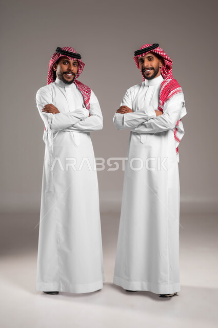 النظر الى الكاميرا بتعابير السرور والثقة بالنفس، مفهوم التوأم الحقيقي المتطابق، بورتريه لشابين توءم عربيين خليجيين سعوديين يرتديان الشماغ والثوب التقليدي يقفان بأيدي متقاطعة، صورة الجسم بالكامل، خلفية رمادية