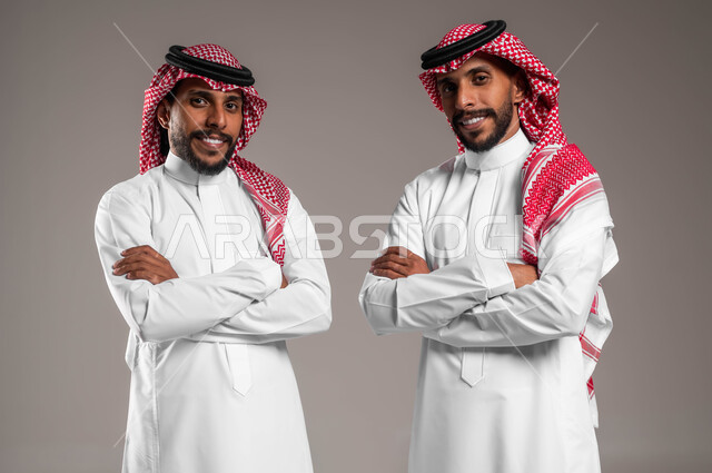 الوقوف باستقامة والنظر إلى الكاميرا بايدي متقاطعة، بورتريه  مقرب لشابين عربيين خليجيين سعوديين يلبسان الثوب السعودي التقليدي والشماغ ينظران باتجاه الكاميرا بإيماءات السرور والسعادة، التعاطف و المودة بين الاخوة، مفهوم العلاقة بين الأخوات، قضاء الوقت مع العائلة، تشارك اللحظات الممتعة، خلفية رمادية