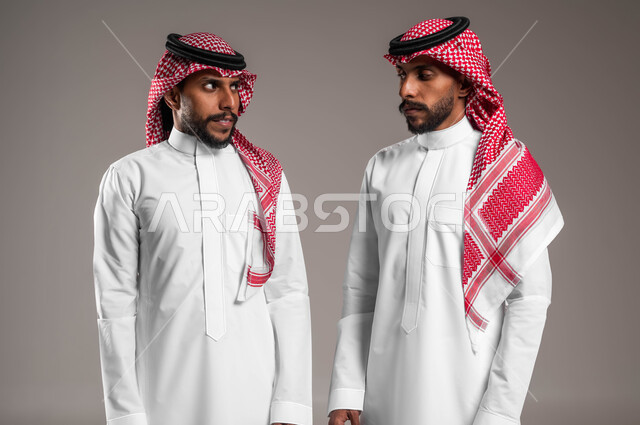 تعابير المواجهة بين الاخوات, النظر الى بعض بثقة, ايماءات التحدي والاصرار, مفهوم لغة الجسد والحركات, بورتريه مقرب من لشابين عربيين خليجيين سعوديين يلبسان الثوب السعودي التقليدي والشماغ يقفان مقابل بعضهما ويتجادلان بإيماءات الغضب وإلقاء اللوم، خلفية رمادية