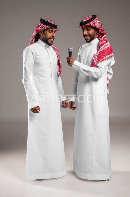 Standing shyly and enjoying singing with the sisters, recording audio clips using the microphone, technical support for singing talents in Saudi Arabia, a portrait of two young Saudi Gulf Arab twins wearing the traditional Saudi dress and shemagh, experimenting with theatrical singing, full body portrait, gray background