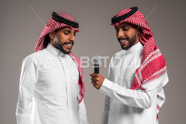 الدعم الفني للمواهب الغنائية في السعودية, الوقوف بخجل والاستمتاع بالغناء مع الأخوات, تسجيل المقاطع الصوتية باستخدام الميكرفون, بورتريه مقرب لشابين توءم عربيين خليجيين سعوديين يلبسان الثوب السعودي التقليدي والشماغ، يقومان بتجربة الغناء المسرحي, خلفية رمادية