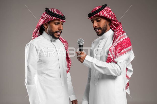 Integration and interaction by singing with sisters, recording audio clips using a microphone, technical support for singing talents in Saudi Arabia, close-up portrait of two young Saudi Gulf Arab twins wearing the traditional Saudi dress and shemagh, experimenting with theatrical singing, full body portrait, gray background