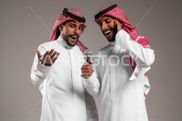 تقديم المواهب الصوتية الفنية الموسيقية بإيماءات الحماس والسرور، الاستمتاع بممارسة هواية الغناء باستخدام الميكرفون, بورتريه مقرب لشابين توءم عربيين خليجيين سعوديين يلبسان الثوب السعودي التقليدي والشماغ، يقومان بعرض غنائي مسرحي, التفاعل والاندماج مع الموسيقى, خلفية رمادية