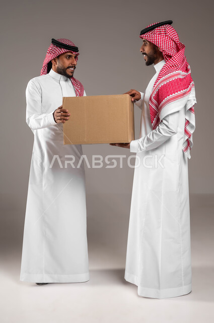 استلام الطلبات الالكترونية، بورتريه لشابين توءم عربيين خليجيين سعوديين، يحملان صندوق مشتريات الاونلاين السريع، جودة شركات الشحن السعودية، خدمات التوصيل المنزلي، التسوق الذكي، صورة الجسم بالطول الكامل، خلفية رمادية