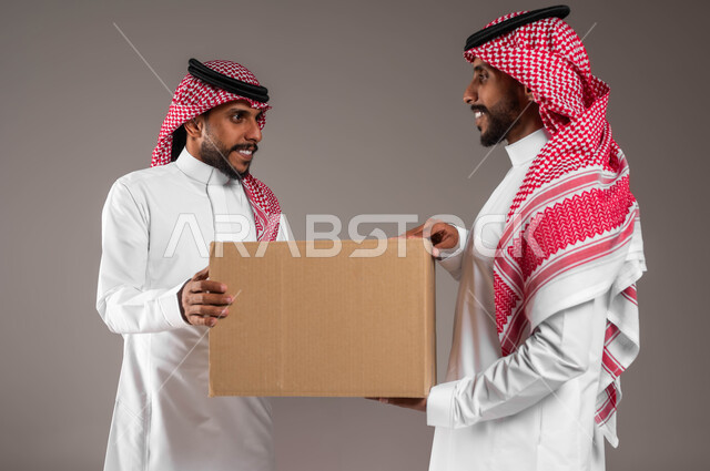 استلام الطلبات الالكترونية، بورتريه مقرب لشابين توءم عربيين خليجيين سعوديين، يحملان صندوق مشتريات الاونلاين السريع، جودة شركات الشحن السعودية، خدمات التوصيل المنزلي، التسوق الذكي، خلفية رمادية