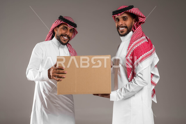 خدمات التوصيل المنزلي، بورتريه مقرب لشابين توءم عربيين خليجيين سعوديين، جودة شركات الشحن السعودية، يحملان صندوق مشتريات الاونلاين السريع، التسوق الذكي، النظر إلى الكاميرا واستلام الطلبات الالكترونية، خلفية رمادية