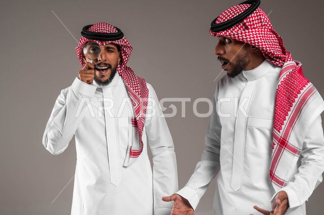 الفحص والاستكشاف والرؤية عن قرب، مفهوم توضيح الصورة، بورتريه مقرب لشاب عربي خليجي سعودي يلبس الثوب السعودي التقليدي والشماغ يمسك بيده عدسة مكبرة برفقة اخاه التوأم, التدقيق والتركيز باستخدام الأدوات البصرية, خلفية رمادية
