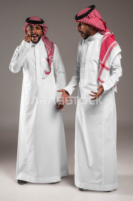 مفهوم توضيح الصورة، بورتريه لشاب  عربي خليجي سعودي يلبس الثوب السعودي التقليدي والشماغ يمسك بيده عدسة مكبرة برفقة اخاه التوأم, الفحص والاستكشاف والرؤية عن قرب, التدقيق والتركيز باستخدام الأدوات البصرية, صورة الجسم بالطول الكامل, خلفية رمادية